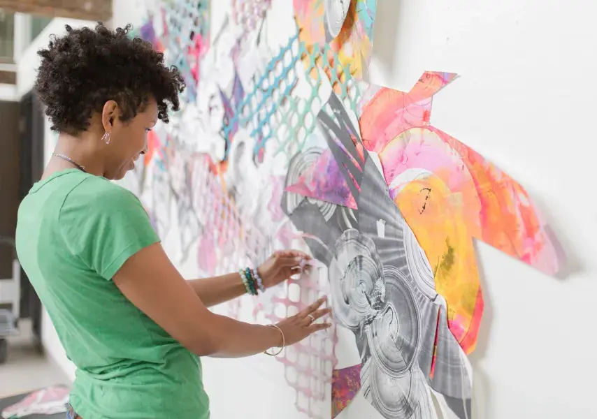 A female artist is captured in her studio, deeply focused on creating a large-scale, abstract wall piece. The artwork is a complex assembly of cut, painted, and printed paper, and she is shown making a delicate adjustment, highlighting the hands-on process of her craft.