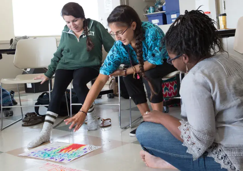 Three participants in an art class or workshop have an informal critique. With artwork displayed on the floor between them, one woman points to a drawing, engaging the other two in a conversation about the pieces.