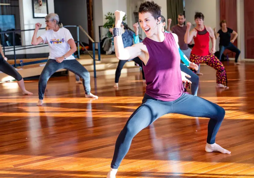 The vibrant energy of a group fitness class fills a sun-drenched studio. A charismatic instructor in the foreground strikes a powerful pose, leading a diverse group of people through an empowering, dance-based workout.