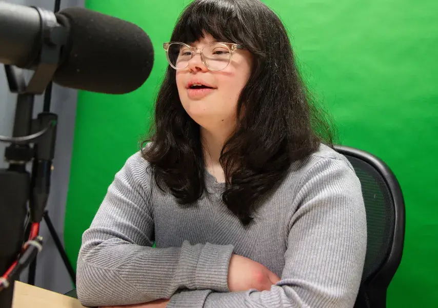 A young woman with long dark hair and glasses is seated at a desk in front of a green screen. She wears a gray ribbed sweater and is speaking into a large black microphone mounted on a stand. Her arms are crossed on the table as she looks slightly upward, suggesting she is engaged in recording a podcast or broadcast in a professional studio setting.