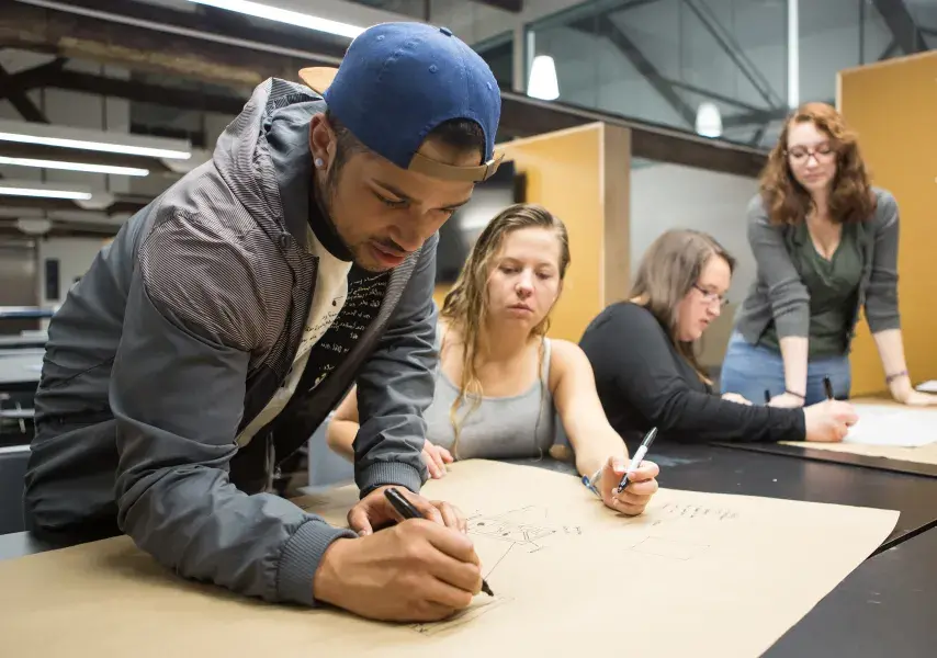 In a design studio, a group of four students gathers around a large table to collaborate on a drawing. A young man in a backwards cap leans over a large sheet of brown paper, actively sketching with a marker, while his three female classmates watch and contribute.