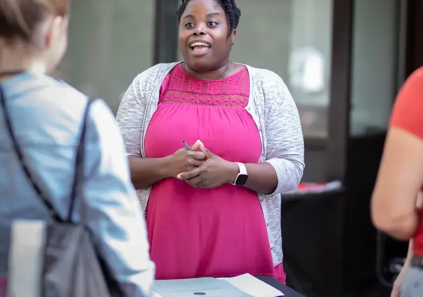 Woman in a pink dress speaking to another student from behind a table