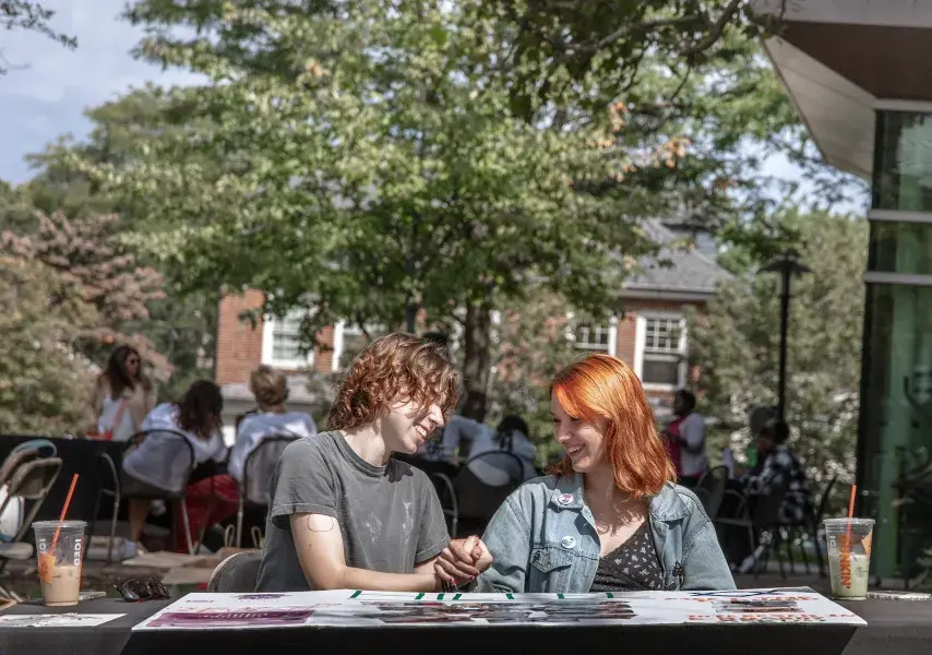 Two students sitting at an outdoor table smiling and holding hands
