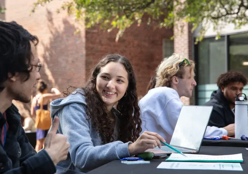 Student with notebook smiling at another person and pointing to a piece of paper with a pencil