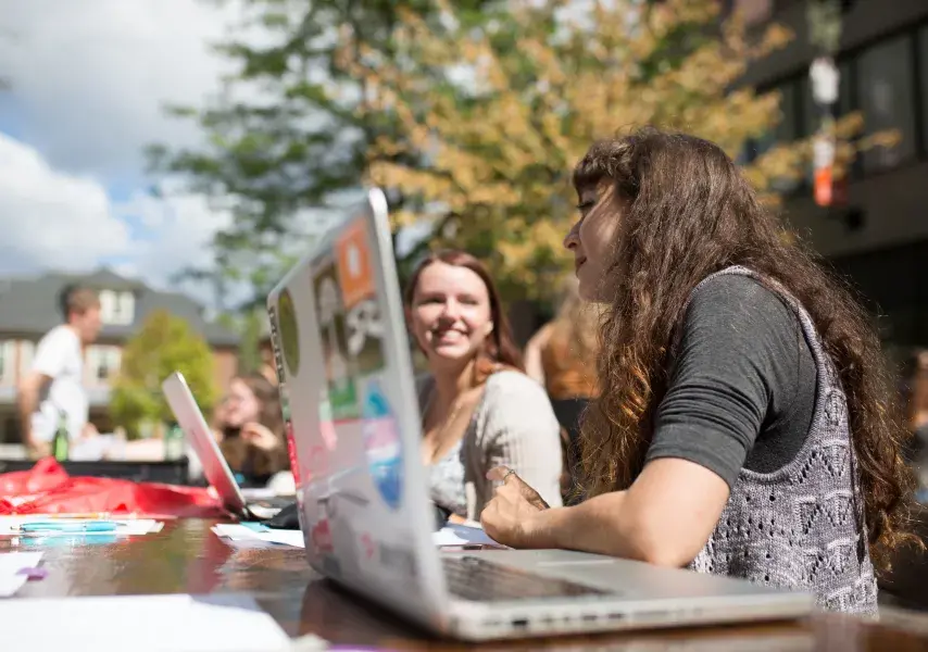 Two students talk to each other behind a laptop at a long outdoor table