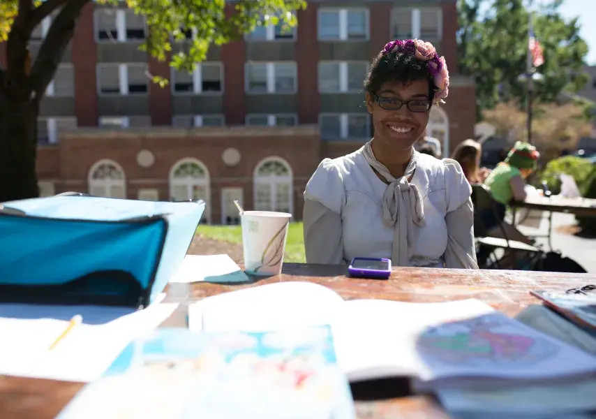 Student smiles behind an outdoor table covered in drawings and notebooks