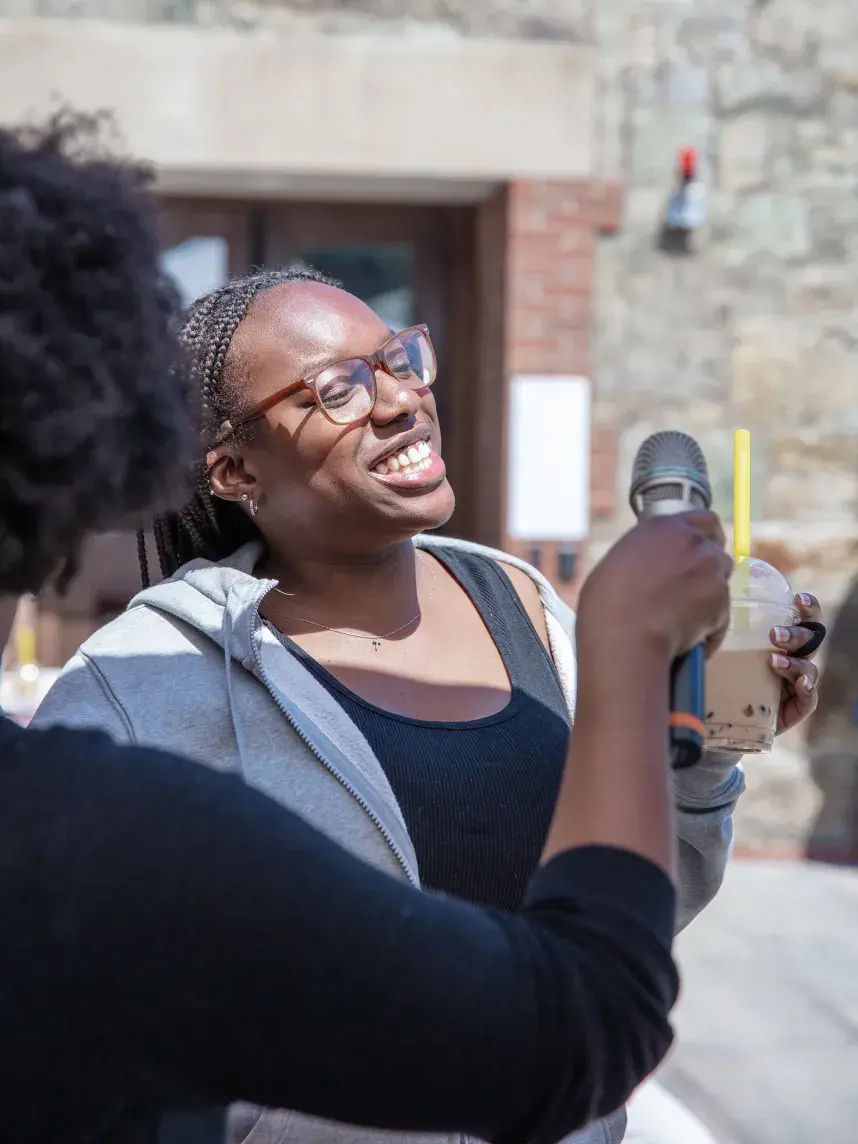 A warm, interactive moment is captured as a young woman smiles during an interview at a wellness fair. She holds a cup of bubble tea while listening to the interviewer, who is visible in the foreground holding a microphone.