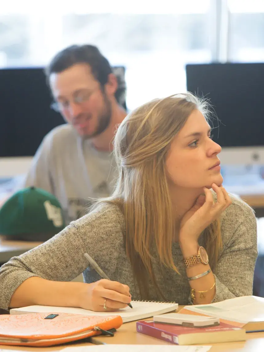 A young woman sits at a table in a classroom, resting her chin on one hand while attentively listening. She has long blonde hair and is wearing a gray sweater. Open books, notebooks, and an orange laptop sleeve are in front of her. In the background, other students are working, and several Apple desktop computers are visible. Natural light streams in through the windows.