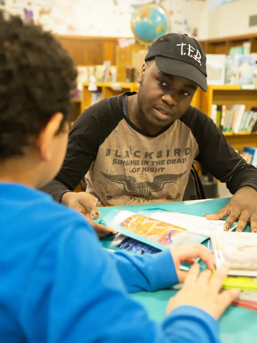 A person wearing a black cap and a shirt with the text "BLACKBIRD SINGING IN THE DEAD OF NIGHT" sits at a table in a library, interacting with a child in a blue shirt. They are looking at colorful, open books spread out on the table, with bookshelves and a globe visible in the background. The setting appears to be an educational or reading activity.
