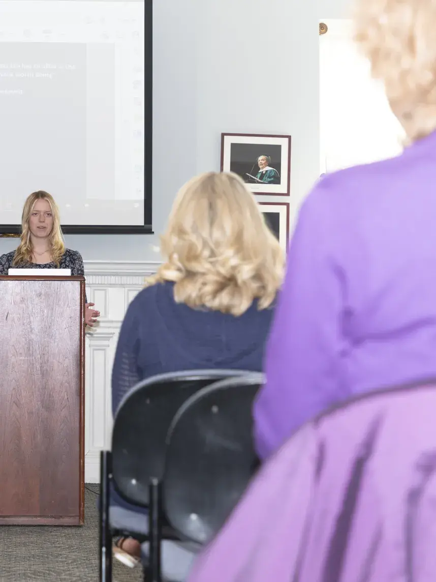A woman stands behind a wooden podium delivering a presentation to an audience seated in rows. A large projection screen behind her displays a slide with an image of a tall building and a quote by Theodore Roosevelt: “Far and away the best prize that life has to offer is the chance to work hard at work worth doing.” Audience members are facing forward, listening attentively. The setting appears to be a formal room, possibly a lecture hall or conference room, with framed portraits on the walls.