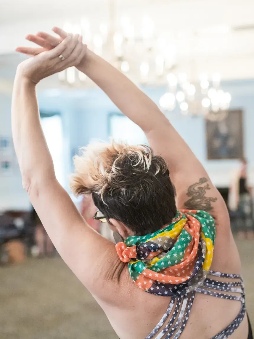 A participant in a mindful movement workshop performs a deep side stretch. The close-up shot from behind focuses on their raised arms and colorful scarf, with the rest of the class and the chandeliers of the room softly blurred in the background.