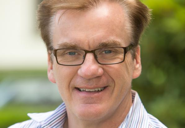 A person with short light hair and glasses in front of greenery
