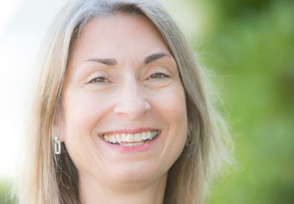 A smiling person with mediu length light hair in front of greenery