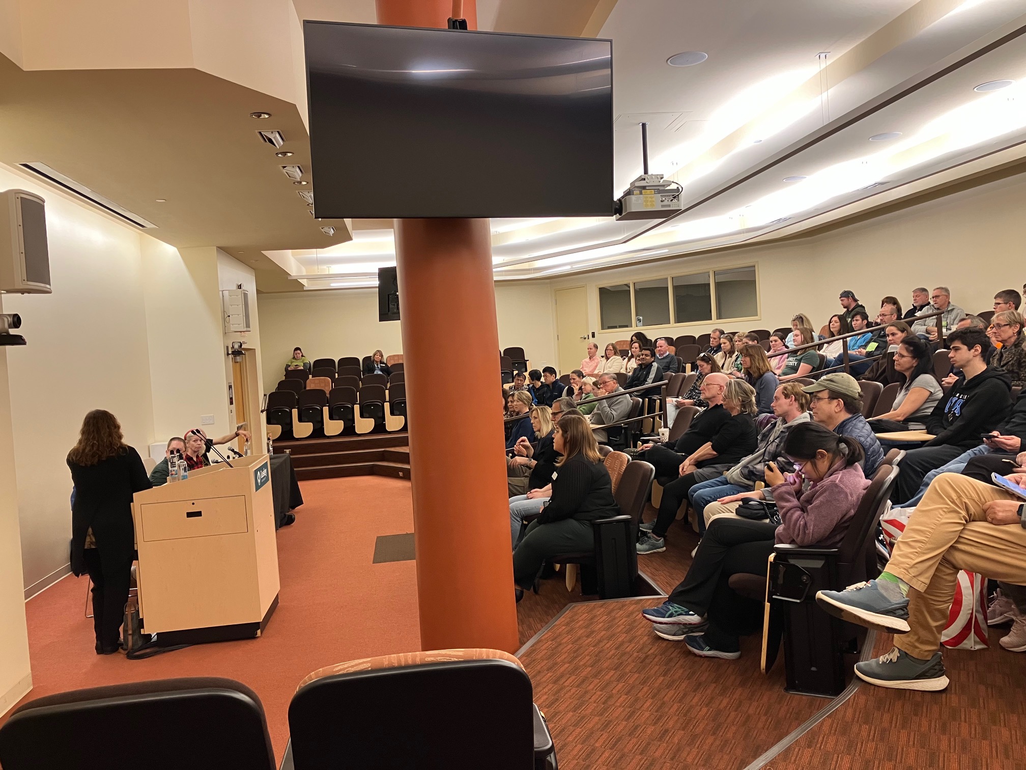 presentation in UHall amphitheater, Jen Thorell at podium, panelists at long table speaking to crowd seated in auditorium